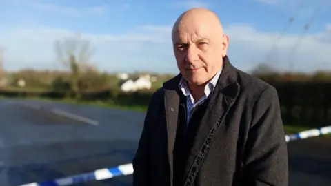 A bald man wearing a black coat over a blue and white striped shirt is standing in front of a rural road which is closed off by a blue and white police cordon. The background is out of focus but a hedgerow, tress and white building can be seen.
