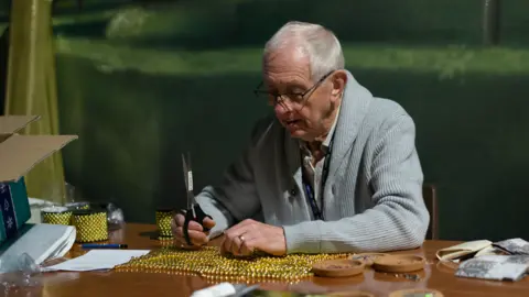 Thursford Christmas Spectacular A man with white hair is wearing a grey cardigan as he sits at a wooden desk holding a pair of scissors as he works with golden-coloured beads and other materials for making theatre costumes.