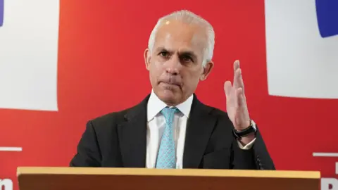 PA Media Ben Habib, leader of Advance UK, wearing a light blue patterned tie, a white shirt and a black suit jacket. He is speaking at a lectern with one hand raised and is standing in front of a large St George's Cross flag