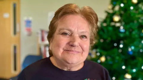 Martin Giles/BBC Sue Hunting has short ginger hair and is wearing a navy jumper. In the background is a Christmas tree which has blue lights and gold baubles on it.