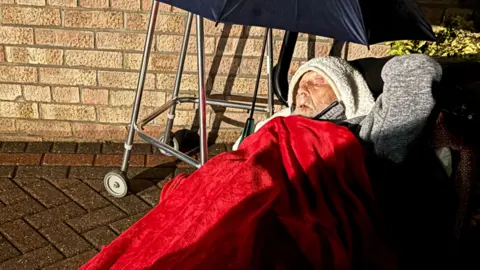 An elderly man lying on a concrete driveway covering in a red blanket and duvets. An umbrella is shielding him from the rain. 