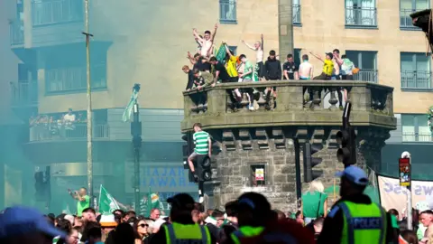 A large gathering of Celtic fans on the Trongate in Glasgow. A number of fans are on top of the Mercat Cross and on traffic lights. Smoke fills the air. The fans are wearing Celtic strips and waving Celtic flags