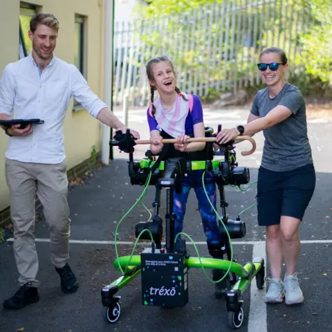 Gwenno Davies Brook in the exoskeleton, she wears a pink and white striped neckerchief and purple top. She is laughing, and walks on a piece of tarmac. A woman wearing sunglasses smiles and holds onto the exoskeleton, while a man holds an ipad and holds the other side. 