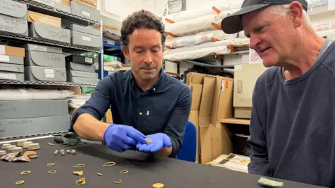 Isles of Scilly Museum Xavier Duffy and Todd Stevens are sitting st a table which is displaying gold rings and coins. Duffy is wearing a pair of blue plastic gloves and is holding an ornate large gold ring, which Stevens is looking carefully at. Behind them are stacked folders and cardboard boxes. Duffy has dark hair and is wearing a black shirt while Stevens has short white hair and is wearing a baseball cap and a blue sweatshirt.