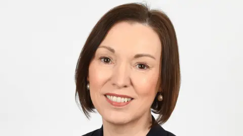 BELFAST TRUST A portrait photo of Jennifer Welsh, who is smiling at the camera and has short brown hair and wearing pearl earrings. She also has black suit jacket on. 