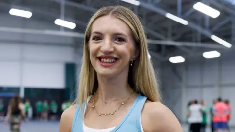 Glasgow 2026 Kira Ewing - a woman with blond hair and a blue top, smiling for the camera during a dance rehearsal inside a sports hall.