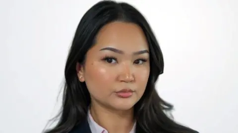 Sunjaya Phillips Sunjaya Phillips has long dark hair and is standing in front of a white background.
