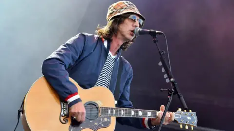 Getty Images Richard Ashcroft performing on stage at Kew The Music 2024 at Kew Gardens, in London on 14 July 2024 in London. He is wearing a blue jacket, black and white striped t-shirt and a tartan brown bucket hat. He is playing the guitar and singing into the mic.