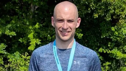 Thomas Mills wears a blue top with a blue ribbon of a medal around his neck. The ribbon says "HALF DRAM 2025 FINISHER". He smiles and stands in front of a tree with lots of green leaves.