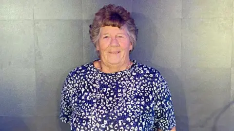 Jo Norman is wearing a navy blue and white patterned short-sleeved top.
She is smiling at the camera and has short brown hair.