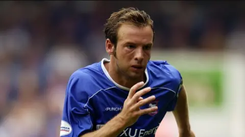 Getty Images Marcus Stewart, pictured in the early 2000s in a head and shoulders image. He is playing for Ipswich Town, wearing their blue kit, with his right arm raised while running. He has short brown hair. 