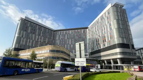 BBC A general view of the hospital, a tall grey building with two wings jutting out from a central structure. There is a blue sky in the background, with buses and green grass in the foreground. 