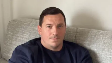 A man sitting on a grey sofa, wearing a blue jumper and a serious expression 