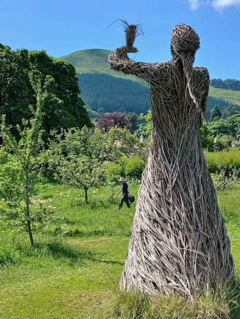 Kenneth Dryburgh The back of a wicker figure looking towards bright green grassy hills and trees on a clear day.