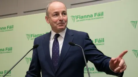 PA Media Micheal Martin who has thinning light coloured hair and is wearing a navy suit and tie with a white shirt looks to his left and points to someone while standing on a podium with Fianna Fail signs in the background