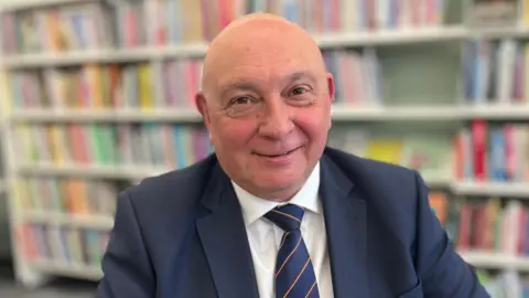 Martin Giles/BBC Carl Smith sitting down in a library looking directly at the camera and smiling. There are rows of books behind him in the background. He is wearing a navy suit with a white shirt and navy striped tie.