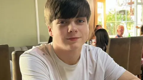 Harry Begg sits at a table in a restaurant, he has short brown hair with a fringe. He wears a white t-shirt