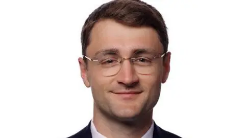 A professional head-and-shoulders portrait of Cai Parry-Jones against a plain light background. He is wearing thin, rimmed eyeglasses with metallic frames.
His hair is short, neatly styled and combed to one side.