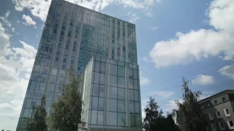 Modern tower with trees in the foreground beneath a bright, partly cloudy sky.