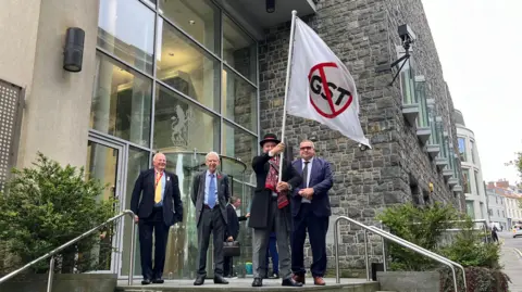 Four men in suits standing in front of Guernsey's Roya Court, the man second from the right wearing a hat is holding a flag which says GSY with a strikethrough. 