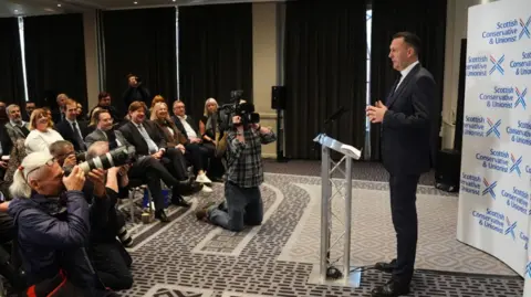 PA Media Findlay stands at a podium in front of photographers