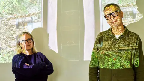 Sally Rodgers and her musical partner Steve Jones are lit by a projector showing distorted bright image. Sally is wearing a purple top and Steve is wearing a green shirt.