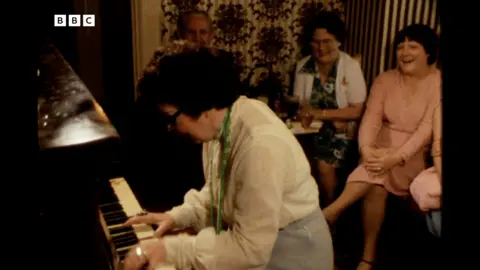 Peggy playing the piano in a pub. Three people are sat behind her watching and laughing.