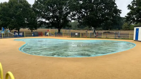 Ben Parker/BBC A drained paddling pool, different coloured mental fences surround the blue pool, which is surrounded by yellow surfacing.
