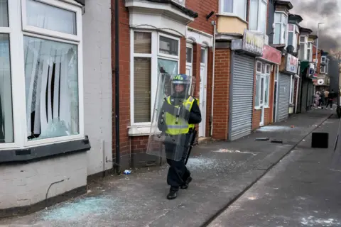 Getty Images A police man holding a riot shield stands in front of a house with smashed windows during a riot
