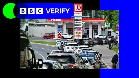 An image of a queue of traffic waiting by a petrol station. It is in a black and blue shared border.