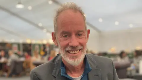 Joanna Durrant/BBC A man with grey hair and beard smiles at the camera. He is wearing a grey jacket and blue shirt 