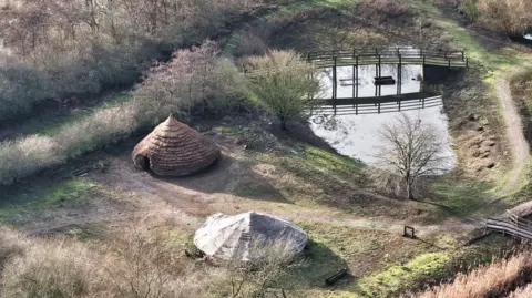 Peterborough From Above Two circular houses made with straw, set in a field with a small water body in the background. The picture was taken from above with a drone.