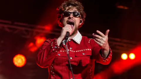 Martin McKay Tom Grennan, on the stage wearing a red jumpsuit, with one arm out and the other holding a microphone. He has short curly hair and is wearing sunglasses. Stage lights are behind him. 