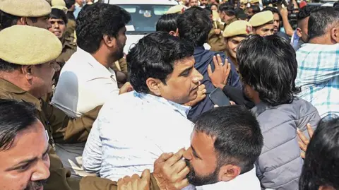 Getty Images Uday Bhanu Chib, wearing a white shirt, surrounded by police officials, walk past a crowd towards a court in Delhi on Tuesday.