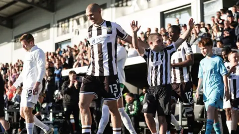 SNS Footballers walk onto a pitch as fans cheer behind them on a sunny day. They are accompanied by mascots. One set of players is wearing black and white striped kits, the other is wearing all white. 