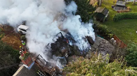 Shaun Whitmore/BBC A drone image above a thatched roof property that is on fire. Thick plumes of smoke billow from the roof. Some firefighters watch on and work from outside the property.