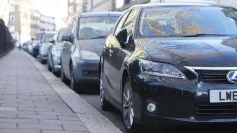 PA Media A close up image of a row of dark-coloured cars parked on a street next to a slab pavement.
