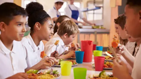 Several primary school pupils sit at a canteen table eating a meal. They are all wearing white uniform polo shirts and are smiling at one another. Alongside the food in front of them are multi-coloured drinking cups and a jug.
