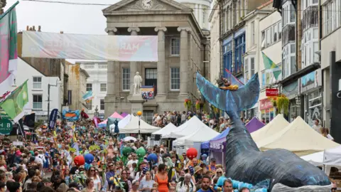 Penzance Council Crowds of people with banners, flags and a giant wicker whale. There are gazebos and shops on the right.