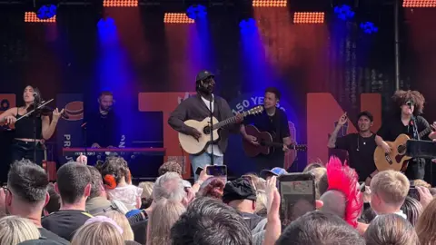 Myles Smith playing a guitar on stage with various other people playing instruments. He is wearing a brown shirt, white T-shirt, jeans and a black baseball cap. A crowd of people are gathered in front of him.