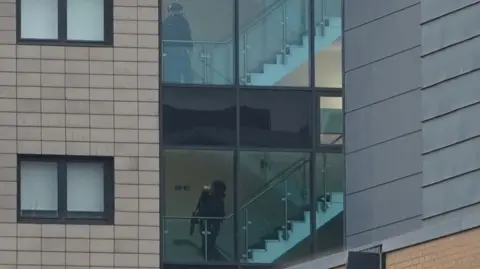 BBC/Oli Constable A modern building with a large glass section that reveals an interior staircase.
Through the tall glass windows, a staircase with white steps and a metal handrail can be clearly seen. Inside the building, two figures dressed in dark clothing and wearing protective helmets are visible on different levels of the staircase. Each figure appears to be carrying a firearm.