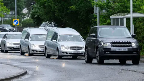 A convoy of silver hearses and mercedes, led by a black Range Rover driving along a road