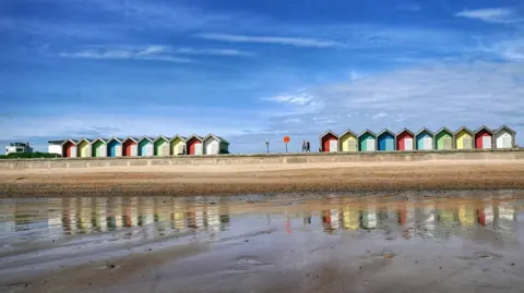 PA Media Wet sand from the tide stretching out into drier sand on a promenade. Colourful beach huts can be seen on the top of a path above the sand. Two people are walking along the path. It is a sunny day.