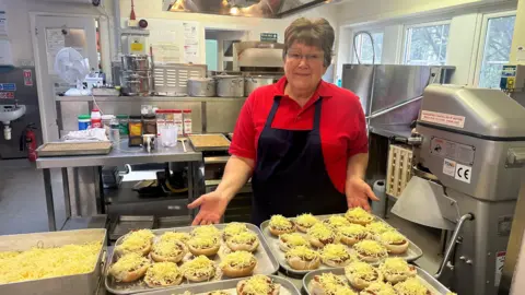 Alison Hopkinson with her hands stretched towards a tray of mini circled pizzas on a baking tray in a school kitchen. 