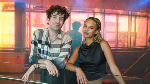 Nick Grimshaw in a checked shirt standing beside Alisha Boe, in black top leaning over banniser