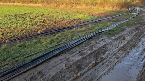 Policing Peterborough A field, that is muddy and wet, with rows of black cable lying on top of it. A blue bucket can be seen in the distance. 