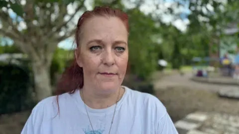 A woman is looking at the camera. She has a somber expression. She's wearing a white T shirt and has dark red hair pulled back in to a ponytail. Behind her is a peaceful garden scene with trees, shrubs and a stone path. 