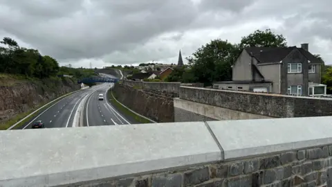 A view from a bridge down onto the Heads of the Valleys dual carriageway. On the right hand side is a large stone wall, and John's house on a platform to the right of the wall. It is a cloudy day.