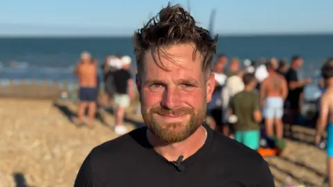 Joe smiles to the camera. He is stood on a pebbly beach with a blue sea behind him and a group gathered on the beach behind him too. It is sunny.