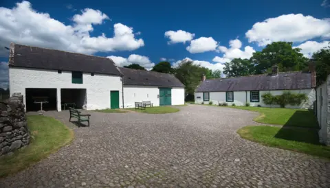 Ian Findlay A number of white farm buildings with green doors and window surrounds, and a cottage, sitting in some gardens with cobbled paths and a few benches